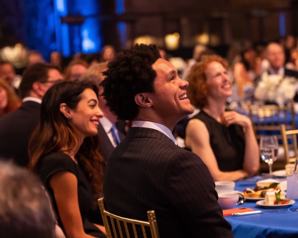 Amal Clooney, Trevor Noah, and Gillian Lester seated at a table, laughing. 