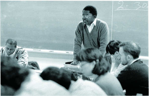 Black and white vintage photo of man in sweater stands at table in front of black board with students