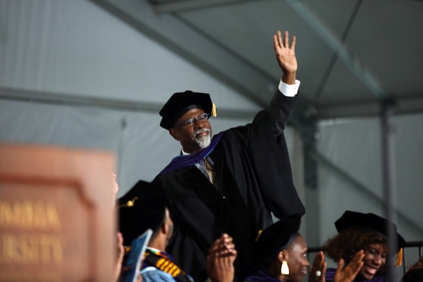 Goateed man in academic regalia raises hand to acknowledge crowd