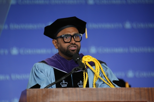 Man with glasses in tam and academic robe speaks at podium