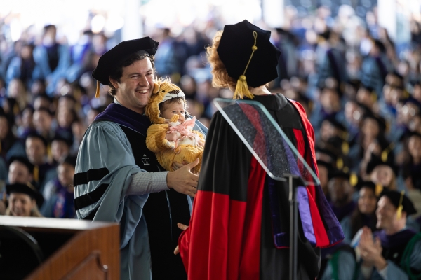 Man in academic regalia, holding baby in lion costume, shakes hands with dean