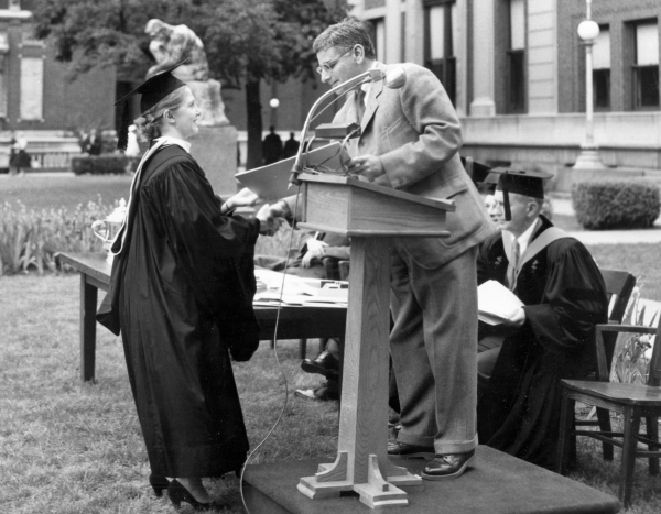A black and white photo showing a woman in a cap and gown receiving an award from a man at a podium