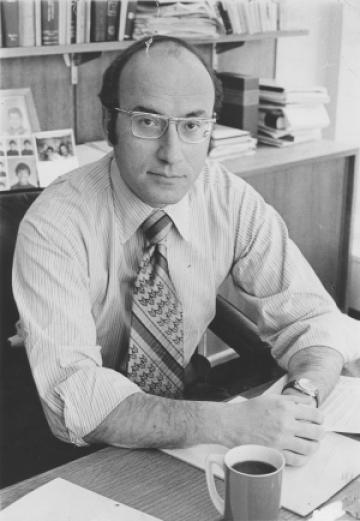 Black and white photo of man with tie sitting at desk with cup of coffee