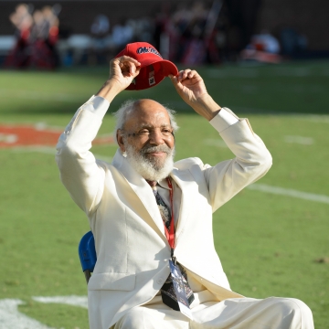 A man in a white suit lifts a red "Ole Miss" baseball cap above his head.