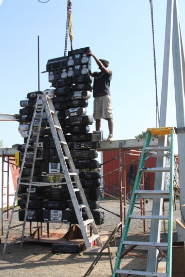 Multimedia artist Bayeté Ross Smith standing on an elevated structure and working on an art installation featuring a tower of radios