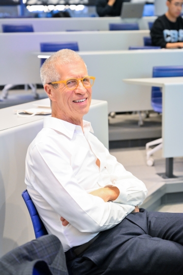 White-haired man in glasses wearing white shirt smiling in classroom