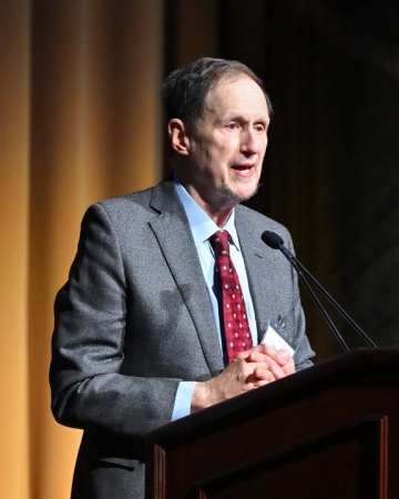 Professor Michael Doyle in light gray suit and red ties talking before a microphone.